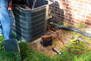 Technician inspecting an outdoor heat pump beside a home while checking wiring and airflow.