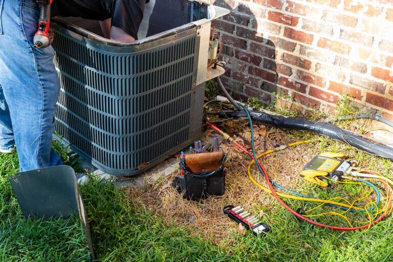 Technician inspecting an outdoor heat pump beside a home while checking wiring and airflow.